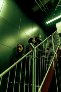 Green photo of band Voice of Baceprot, posing on stairwell under fluorescent lighting